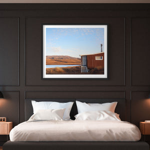 Framed landscape print of a Lake Onslow fishing hut above a bed in a bedroom with dark paneling.