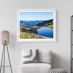 Framed landscape art print of a scene of a mountain tarn on the St Arnaud range at the nelson lakes on a wall above a sofa.
