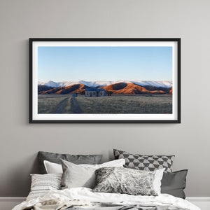 Framed landscape art print of the Hawkdun range mountains in Otago behind a field and hut above a bed with pillows.